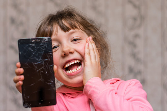 Portrait of a cheerful little girl with a mobile phone. The child broke the telephone communicator and laughs. Baby joked breaking smartphone screen.