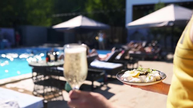 Waiter carries oysters and scallops on tray and glass of champagne or a sparkling wine near swimming pool at luxury hotel or beach club. Man serving seafood and drinks in slow motion at resort.