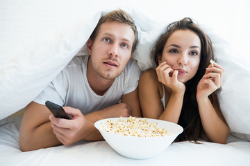 young couple handsome man and beautiful woman watching TV in bed eating pop corn looking fascinated
