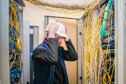 A sad engineer in a white helmet took hold of his head in his hands, taking a wrong decision. Incorrect connection of communication equipment in the data center server room.