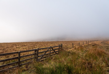 A wooden fence that stretches between Scotland and England border with brown grass field on either side on a foggy and grey day.