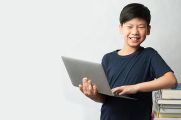 Online learning concept. A smart looking Asian boy standing with a computer laptop and a tall stack of colorful and various types of text books beside him. E-learning, Gamification, Self- Study.  © myboys.me