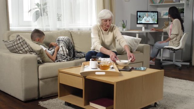 Wide shot of grandmother with grey hair putting cold compress on forehead of sick boy as young mother looking at computer screen and giving her instructions from online doctor
