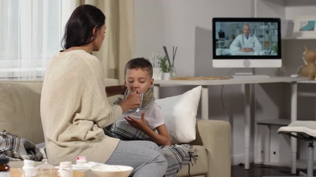 Medium shot of caring young mother talking on video call with online doctor and giving water to sick little boy lying on sofa and playing on tablet