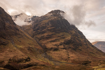 One of the three mountains called The Three Sisters, covered in brown wintery vegetation on a partially cloudy day in Scottish Highlands.