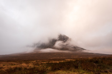 Wintery brown grass fields overlooks Buachaille Etive Mòr mountain covered in clouds in the Scottish Highlands.