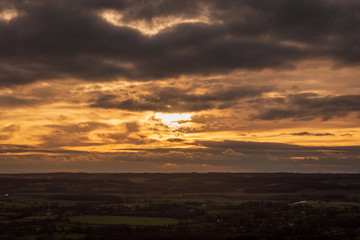 The sun sets through dramatic clouds that loom over horizon overlooking the English countryside.