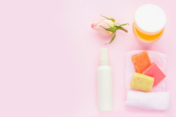 Open jar of cosmetic cream, white cosmetics bottle, soap, rose and towel on pink background. Concept of natural spa cosmetics. Flat lay, top view, copy space .
