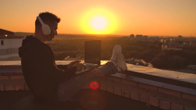 A programmer with a laptop in headphones sits on the roof and writes code at sunset and listens to music. Remote work freelancer. Freedom to work, Typing on keyboard at sunset with a view of the city