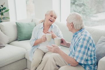 Photo of two pretty aged people excited pair laughter communicating tea mugs hands comfortable flat sitting sofa indoors