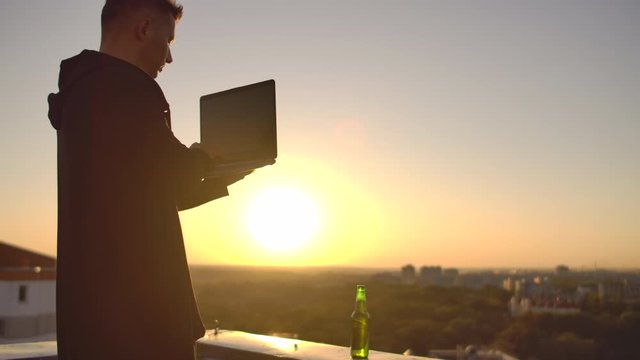 A programmer with a laptop sits on the roof and writes code at sunset. Remote work freelancer. Freedom to work. Typing on a keyboard at sunset with a view of the city