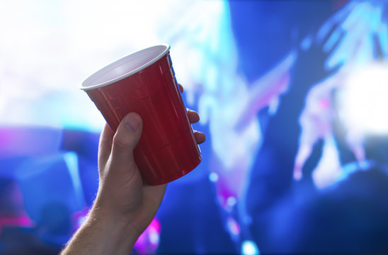 Young man holding red party cup in nightclub dance floor. Alcohol container in hand in disco. College student having fun and dancing. Celebrating people in the back.