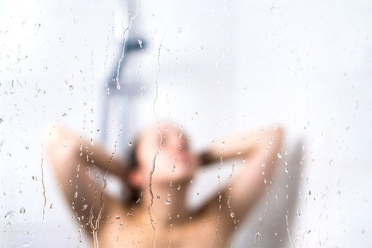 Young beautiful woman in shower. Behind wet glass window with water drops. Focus on droplets. Girl bathing, washing hair and taking fresh bath.