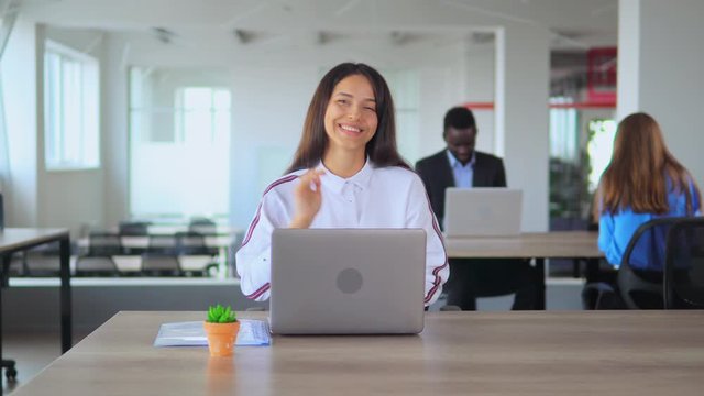 attractive girl office employee looking camera smiling showing hand gesture hello at workplace cheerful business woman in casual white shirt sitting at desk