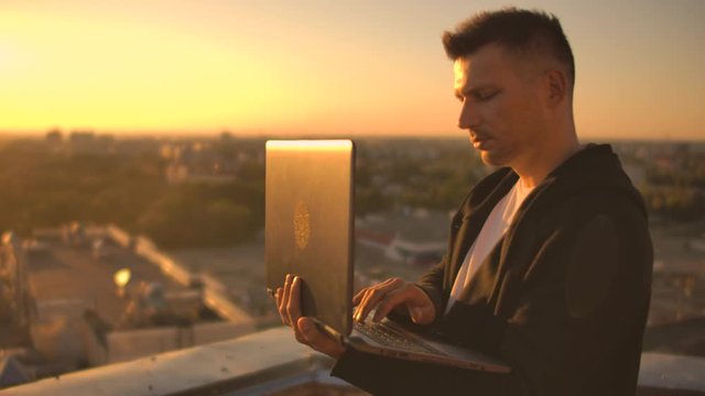 A programmer with a laptop sits on the roof and writes code at sunset. Remote work freelancer. Freedom to work. Typing on a keyboard at sunset with a view of the city