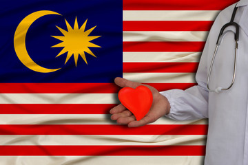 photo of a doctor with a stethoscope with a heart in his hand against the background of the national flag of state of Malaysia, the concept of health care, cardiological treatment, medical insurance