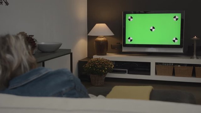Wide shot of a young woman watching TV in a modern living room with green screen chroma key options for VFX