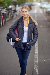 Portrait of young blond woman in city street, holding hands in her casual black unzipped jacket, looking at camera and smiling