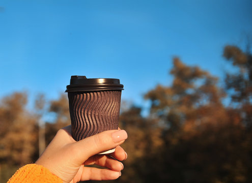 Female hand with brown paper cup of coffee on autumn background.