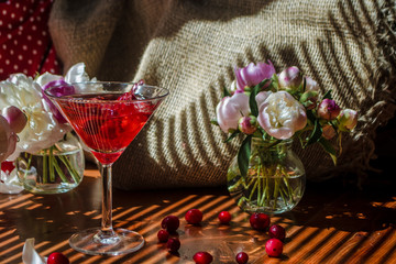 Solemn still life with glass of red drink with autumn berries with a splash and splatters of falling berry surrounded by white flowers on textile background in diagonal shadow. Sunset in rich colors