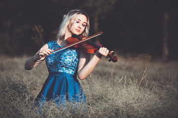 portrait of a young fabulously beautiful girl in a dress with a violin sitting in dry grass on meadoe at the dawn, woman playing a musical instrument with inspiration relaxing on nature © fantom_rd