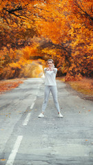 Fototapeta premium slim young woman in sportswear exercises and stretches hands up satanding on asphalt road, girl engaged in sport outdoors on background of autumn foliage, concept healthy lifestyle and female beauty