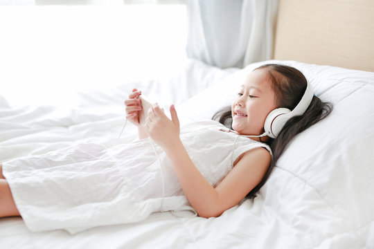 Happy little Asian girl using headphones listen music by smartphone smiling while lying on bed at home.