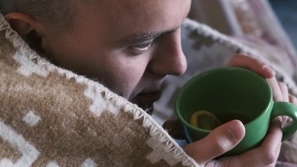Unhealthy young man drinking hot tea wrapped in a plaid, close-up