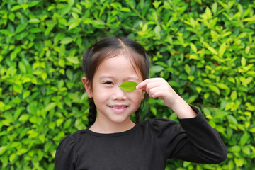Portrait of smiling little Asian child girl holding a green leaf closing right eye in green garden background.