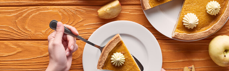cropped view of woman holding spatula with piece of pumpkin pie with whipped cream near cut and whole apples on orange wooden table, panoramic shot