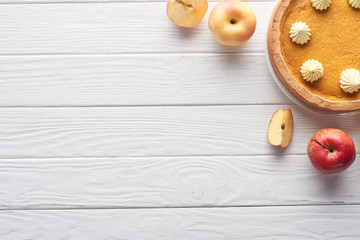 top view of delicious pumpkin pie with whipped cream near cut and whole apples on white wooden table