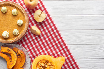 tasty pumpkin pie with whipped cream on checkered napkin near raw and sliced baked pumpkins, cut and whole apples on white wooden table