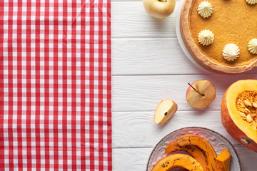tasty pumpkin pie with whipped cream near checkered tablecloth, raw and baked pumpkins, whole and cut apples on white wooden table