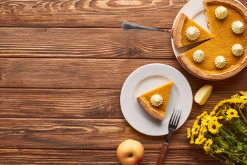 cut pumpkin pie with whipped cream, apple, and yellow flowers on wooden surface