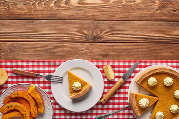 cut pumpkin pie with whipped near baked sliced pumpkin, fork and knife of checkered tablecloth on wooden surface