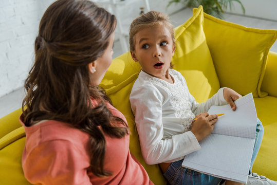 Surprised Kid Holding Notebook And Looking At Babysitter In Living Room