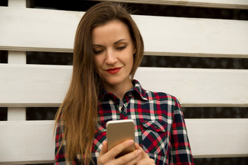 Young beautiful girl stands near a white wall, smiles and holds a mobile phone, looks at the phone screen.