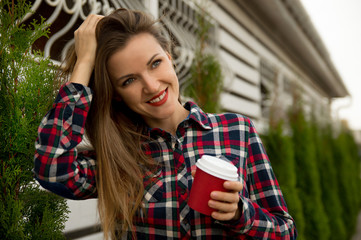 Young beautiful girl is walking down the street, smiling and holding a red paper cup with coffee.