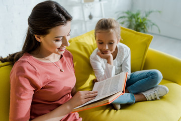 selective focus of happy kid sitting on sofa with happy babysitter reading book