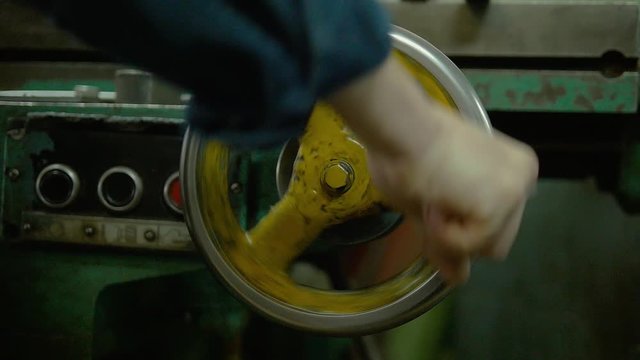 A factory worker is spinning with his fist a metal wheel of some appliance on a plant. We can see a lot of gear and heavy machinery around.