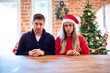 Young couple wearing santa claus hat sitting on chair and table around christmas tree at home...