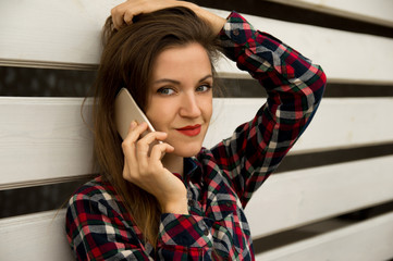 A young beautiful girl is standing near a white wall and talking on a cell phone.