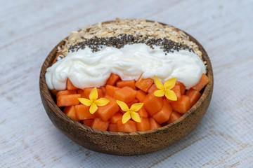Slices of sweet papaya with oat flakes, chia seeds and white yogurt in coconut bowl on white wooden background, close up