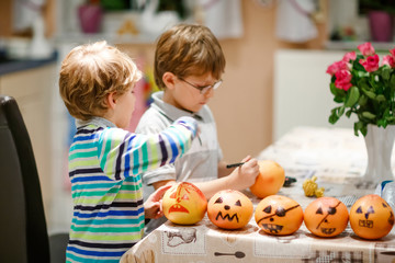 Fototapeta premium Two little kids boys making Jack-o-lantern with drawing scary faces on mandarine, tangerine or clementine. Happy children siblings and twins making preparation for Halloween party at home, indoors