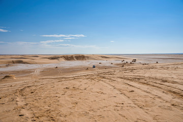 Sahara desert, with a salt marsh plot, with sand dunes, traces of past cars, and an off-road vehicle moving in the background