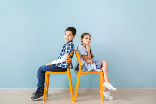 Cute Fashionable Children Sitting On Chairs Near Color Wall