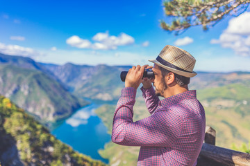 Fashionable man using binoculars and looking at beautiful nature with mountains and canyon.