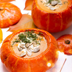Fresh creamy orange pumpkin soup in baked pumpkins with green dill and pumpkin seeds on white wooden background with yellow and red autumn leaves and bokeh lights. Top view