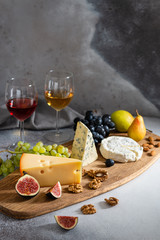 Delicious cheeses served on a wooden board with fruits and nuts and two glasses of red and white wine on gray background. Copy space, side view. Wine and cheese