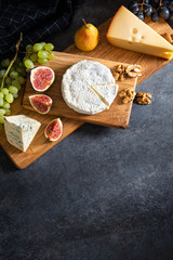 Cheese plate: Maasdam, Camembert cheese, blue cheese, figs, walnuts, almonds, pears and grapes on wooden board on gray background. Copy space for text. Top view, falt lay. Appetizer, menu